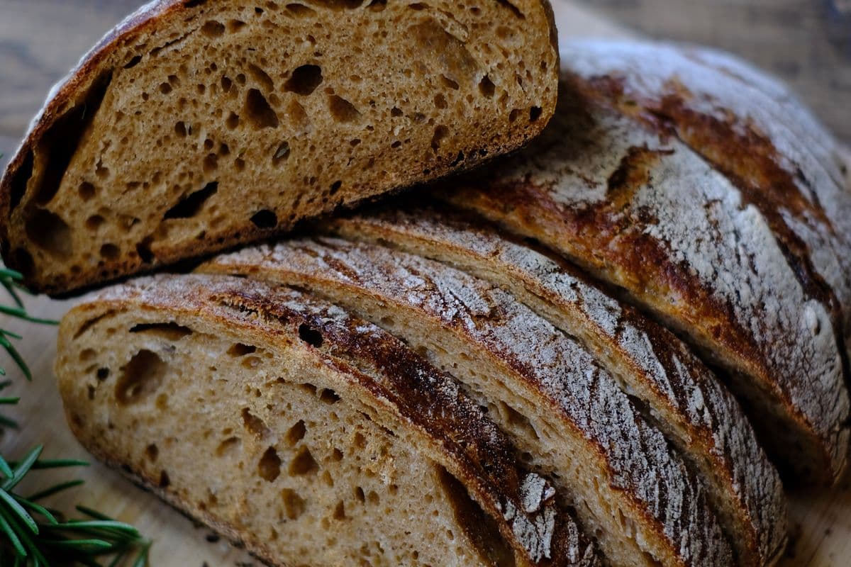 Sliced crusty loaf of freshly baked bread showing its open crumb, resting on a wooden board with a sprig of rosemary
