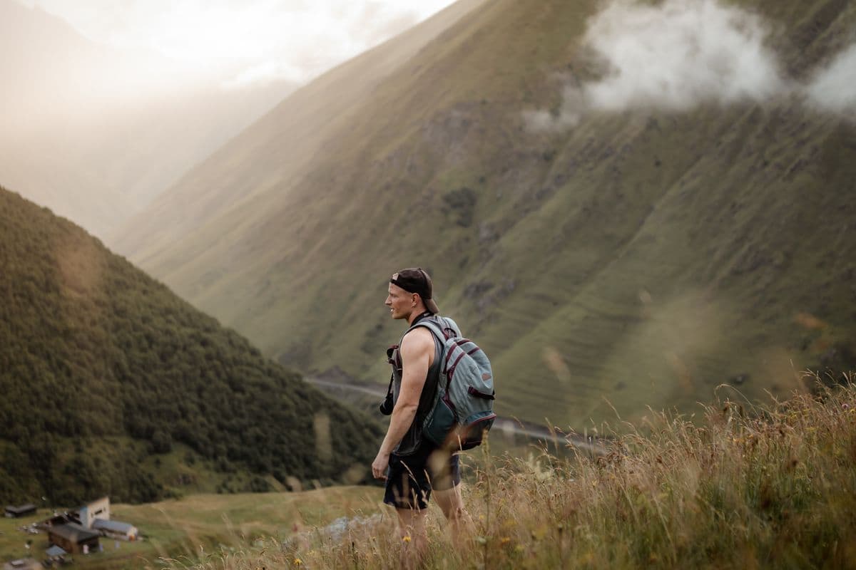 Hiker wearing a daypack walking through tall grass on a mountainside with misty ridges behind