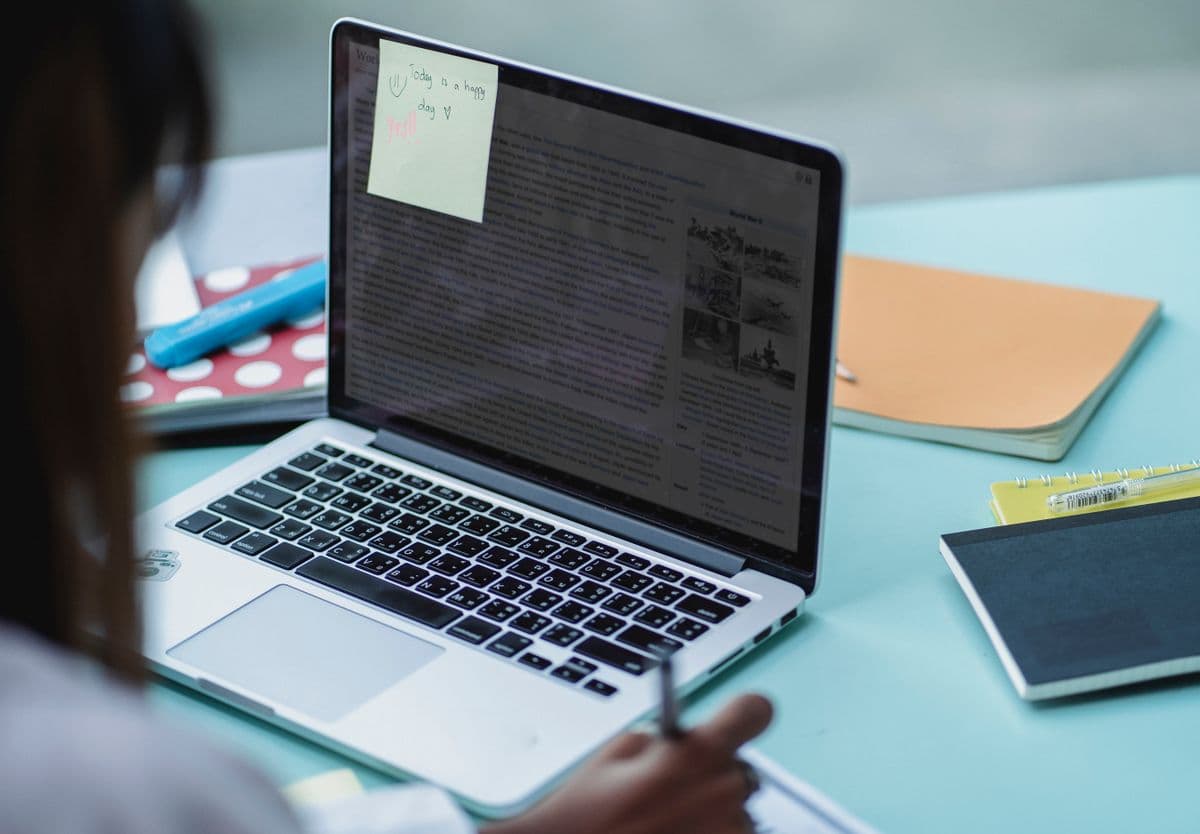 Student taking notes at a table in front of an open laptop with notebooks alongside