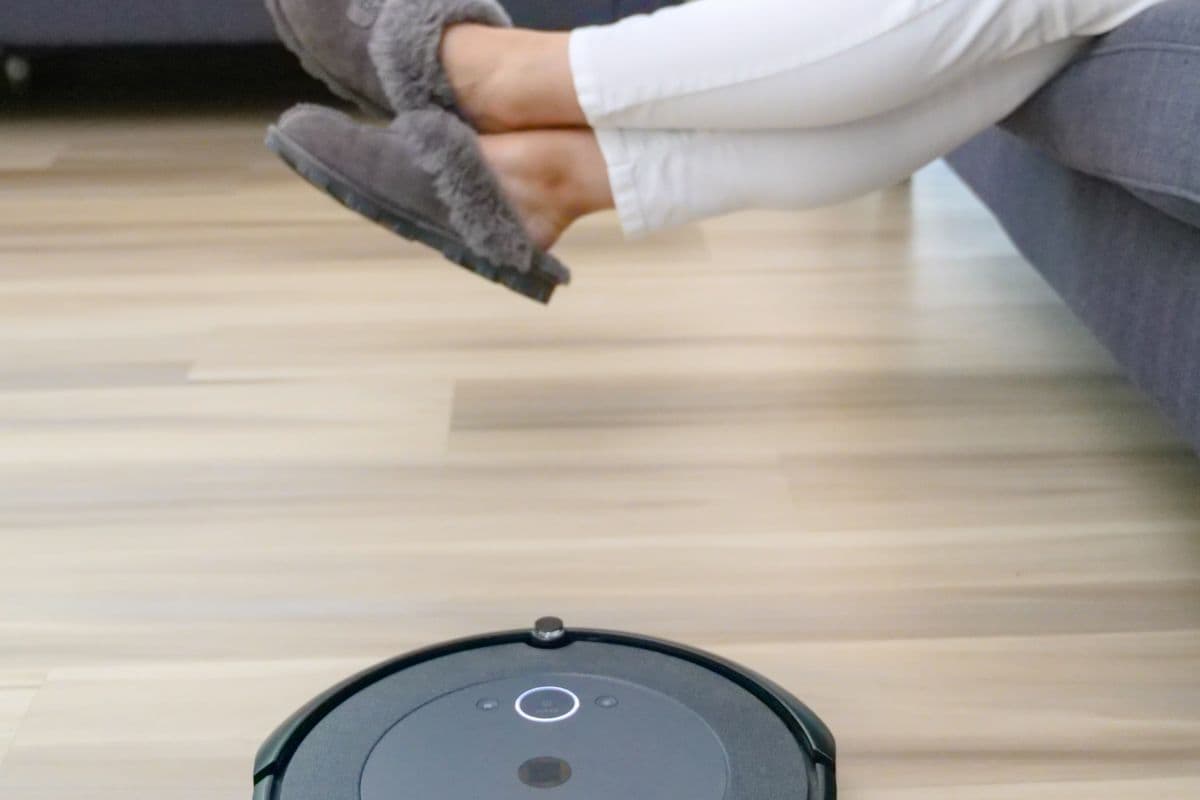 Round dark robot vacuum cleaning a light hardwood floor while a person rests on a couch behind it