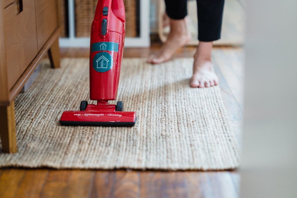 Red lightweight stick vacuum standing on a woven rug in a living room with a person walking behind