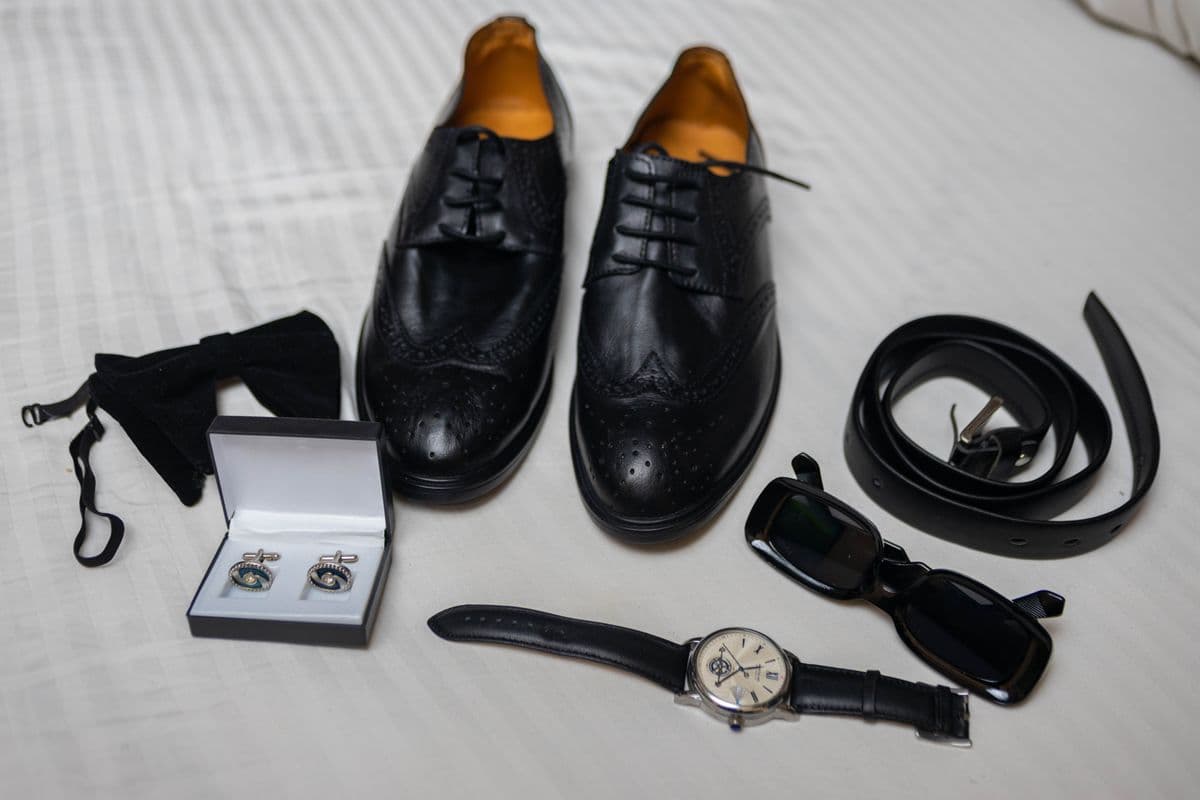 Flat lay of black leather work accessories on a bed: dress shoes, a belt, a wristwatch, sunglasses, cufflinks, and a bow tie